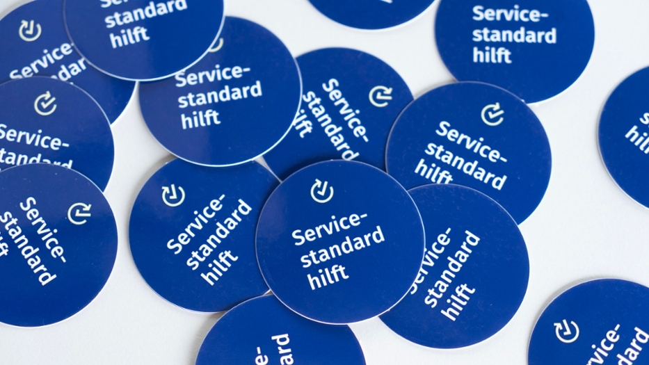 Größere Menge von blauen, runden Aufklebern mit der Aufschrift "Servicestandard hilft", die auf einer weißen Tischplatte liegen.