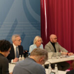 Karsten Wildberger, Kirsten Sinemus und Fabian Mehring sowie mehrere unbekannte Personen in Rückenansicht in einem Sitzungssaal.