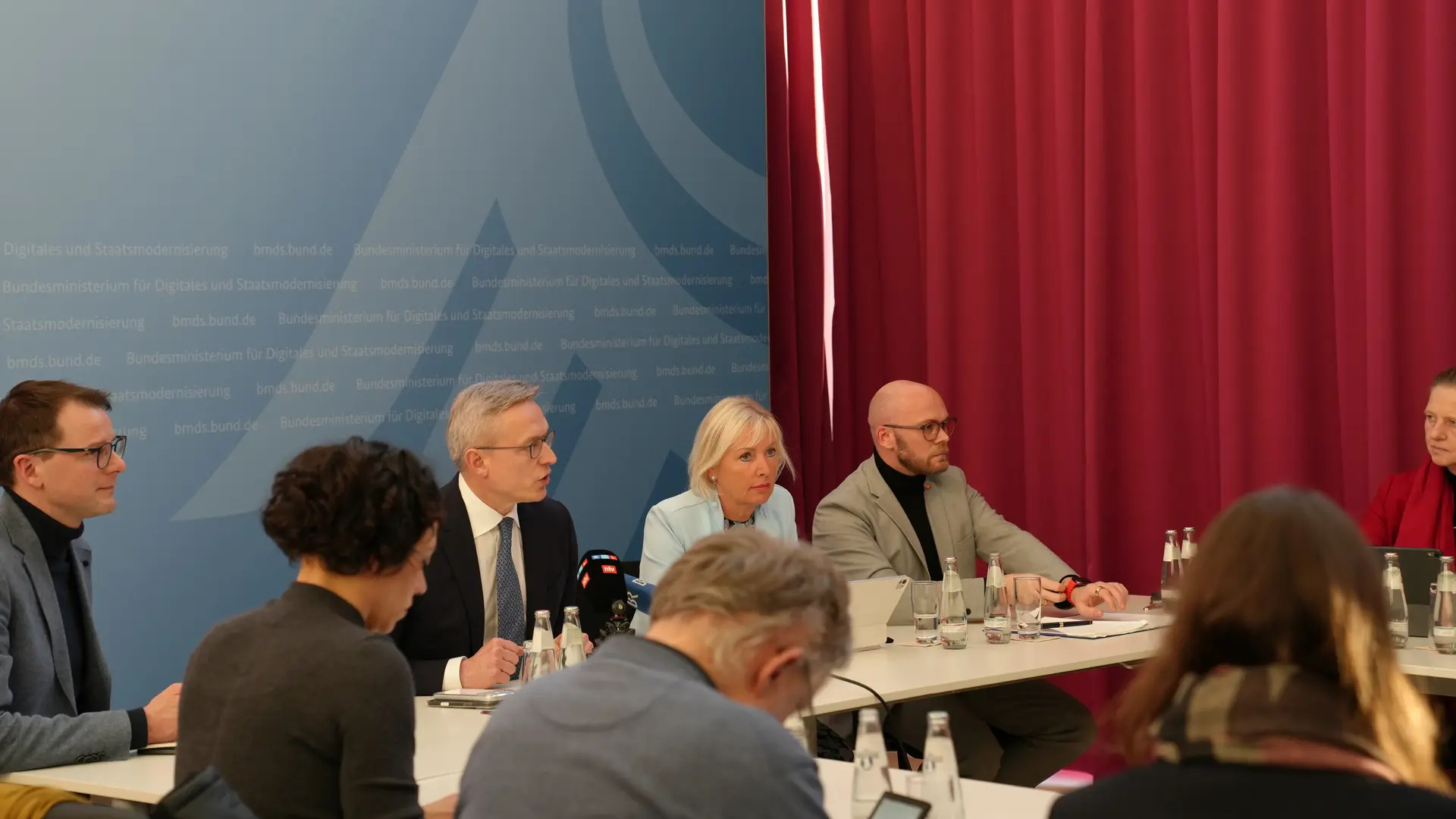 Karsten Wildberger, Kirsten Sinemus und Fabian Mehring sowie mehrere unbekannte Personen in Rückenansicht in einem Sitzungssaal.