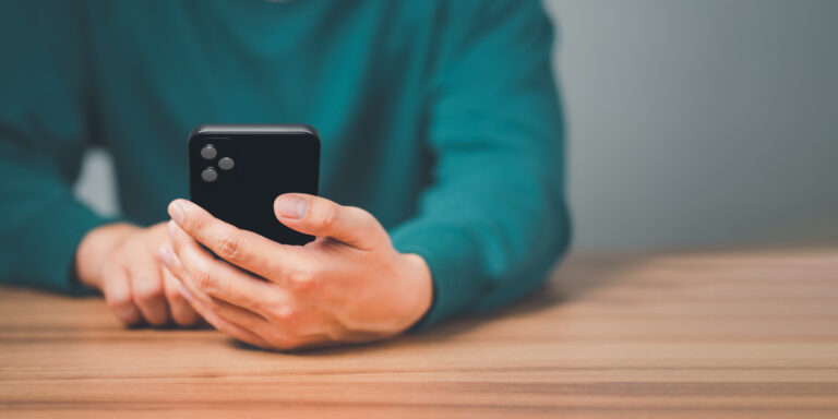 Nahaufnahme einer Person (Torso) mit grünem Pullover, die an einem Holztisch sitzt und mit dem Smartphone hantiert.