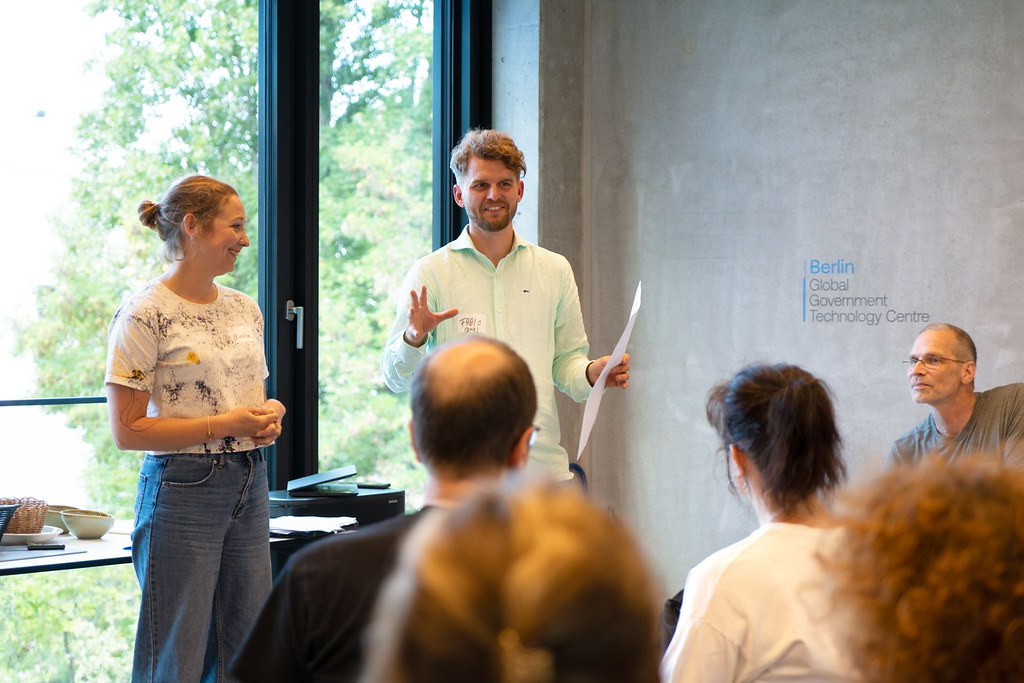 Zwei Personen stehen vor einem Fenster und präsentieren etwas. Im Vordergrund sitzen einige Personen schauen sie an und hören zu.