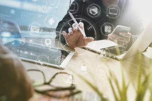 Montage: Schreibtisch, Laptops, Hände von Jacketttragenden Männern, Überlagerung mit diversen IT-Icons