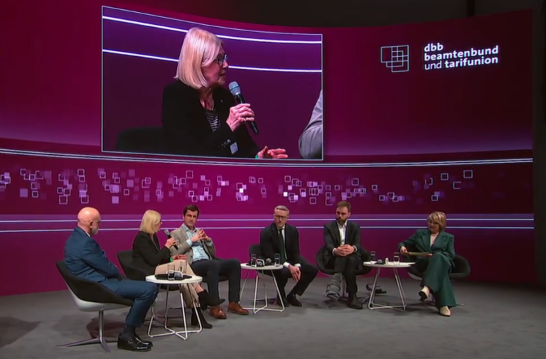 Purpurfarben ausgeleuchtetes Podium mit Diskususionsteilnehmenden auf Sesseln