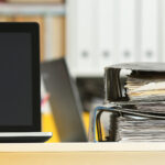 Leeres Büro, Nahaufnahme eines Schreibtischs, auf dem ein Laptop und abgewetzte Aktenordner nebeneinander liegen. Im Hintergrund weitere Akten in Regalen.