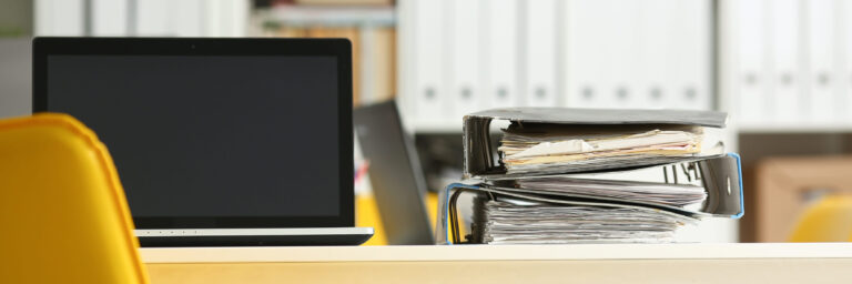 Leeres Büro, Nahaufnahme eines Schreibtischs, auf dem ein Laptop und abgewetzte Aktenordner nebeneinander liegen. Im Hintergrund weitere Akten in Regalen.
