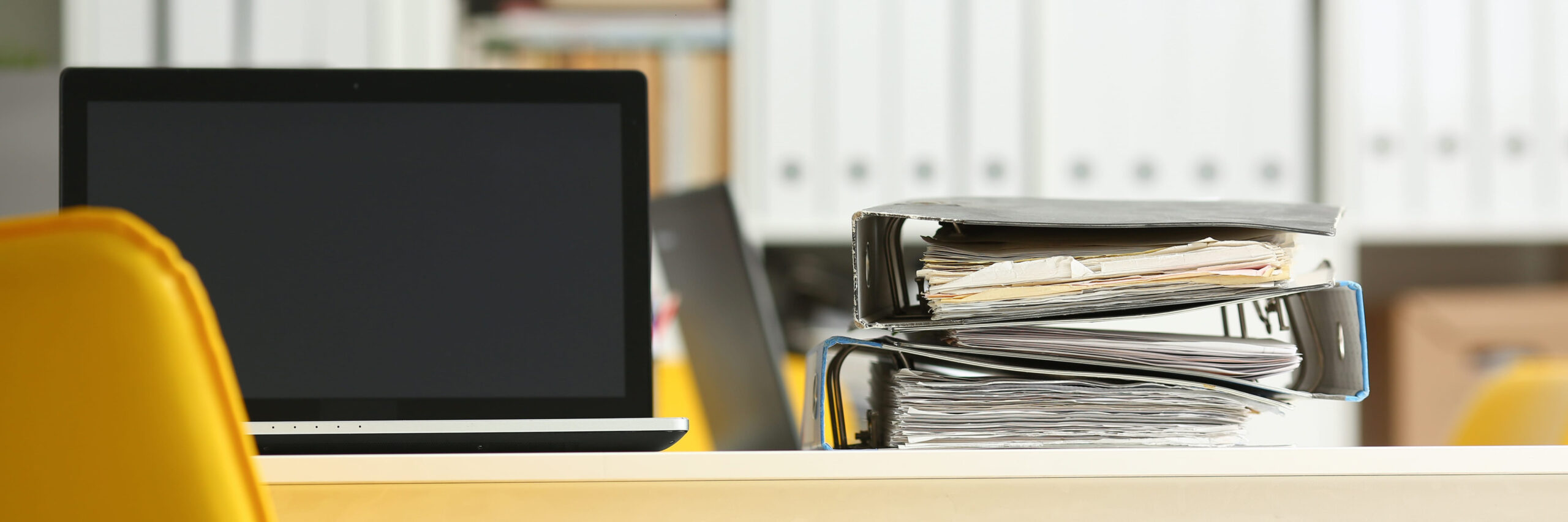 Leeres Büro, Nahaufnahme eines Schreibtischs, auf dem ein Laptop und abgewetzte Aktenordner nebeneinander liegen. Im Hintergrund weitere Akten in Regalen.