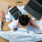 Blick von oben auf einen Schreibtisch mit Laptop, Ordner und papieren. Ein Mann im weißen Businesshemd ist über seiner Arbeit zusammengebrochen.