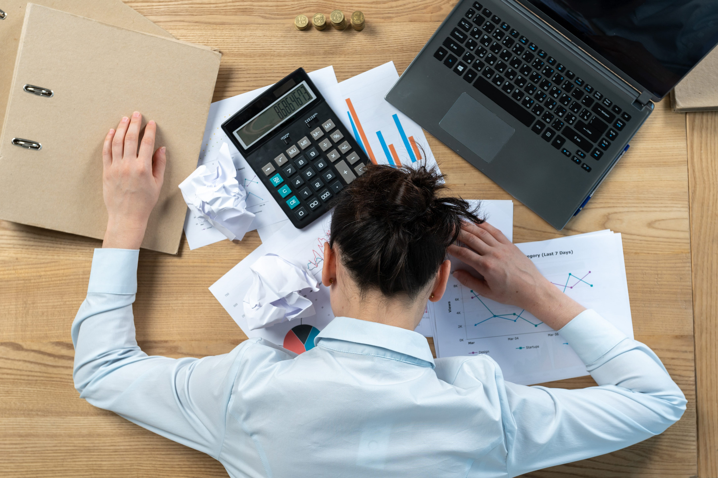 Blick von oben auf einen Schreibtisch mit Laptop, Ordner und papieren. Ein Mann im weißen Businesshemd ist über seiner Arbeit zusammengebrochen.