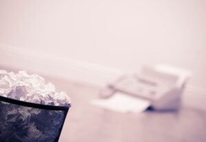 Papierkorb mit zusammengeknülten Papieren, im Hintergrund unscharf ein Fax-Gerät