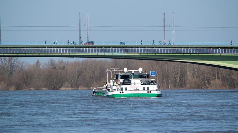 Binnenschiff fährt unter einer Brücke mit Solarpanelen hindurch.
