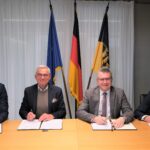 Vier Männer in dunklen Anzügen sitzen nebeneinander an einem Holztisch, vor sich Dokumente.