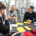 Mehrere Personen sitzen offenbar miteinander redend um einen Tisch herum, dieser ist mit gelben Zetteln bedeckt.