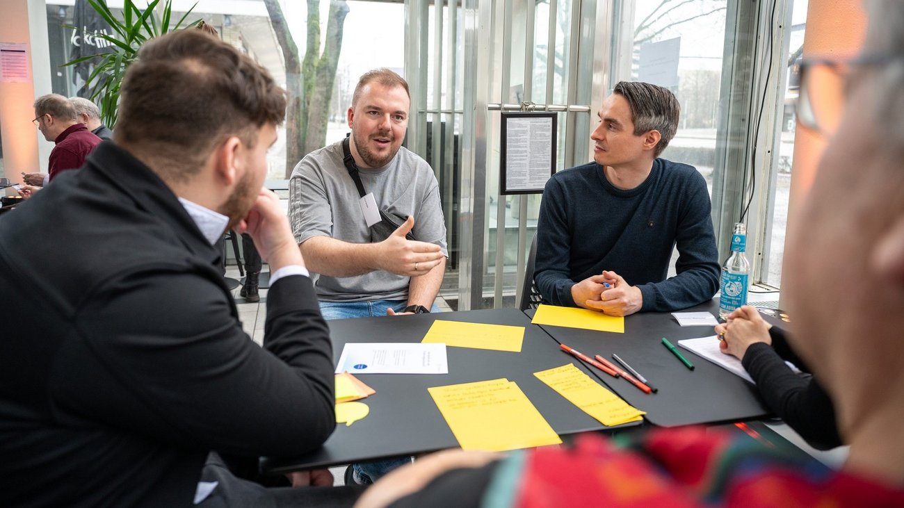 Mehrere Personen sitzen offenbar miteinander redend um einen Tisch herum, dieser ist mit gelben Zetteln bedeckt.