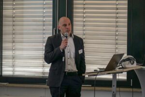 Kahler Mann im Anzug mit Mikro in der Hand an einem Rednerpult, im Hintergrund zugezogene Jalousien.