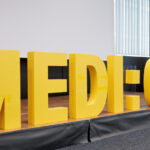 Bühne mit Blumenbukett, im Vordergrund sind die dreidimensionalen, gelben Majuskeln "MEDI:CUS" aufgestellt