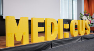 Bühne mit Blumenbukett, im Vordergrund sind die dreidimensionalen, gelben Majuskeln "MEDI:CUS" aufgestellt