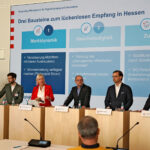 Sechs in einer Reihe stehende Rednerpulte, alle bis auf eines mit dunkel gekleideten Männern besetzt, am 3. von links Kristina Sinemus in leuchtend rotem Blazer.