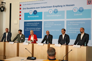 Sechs in einer Reihe stehende Rednerpulte, alle bis auf eines mit dunkel gekleideten Männern besetzt, am 3. von links Kristina Sinemus in leuchtend rotem Blazer.