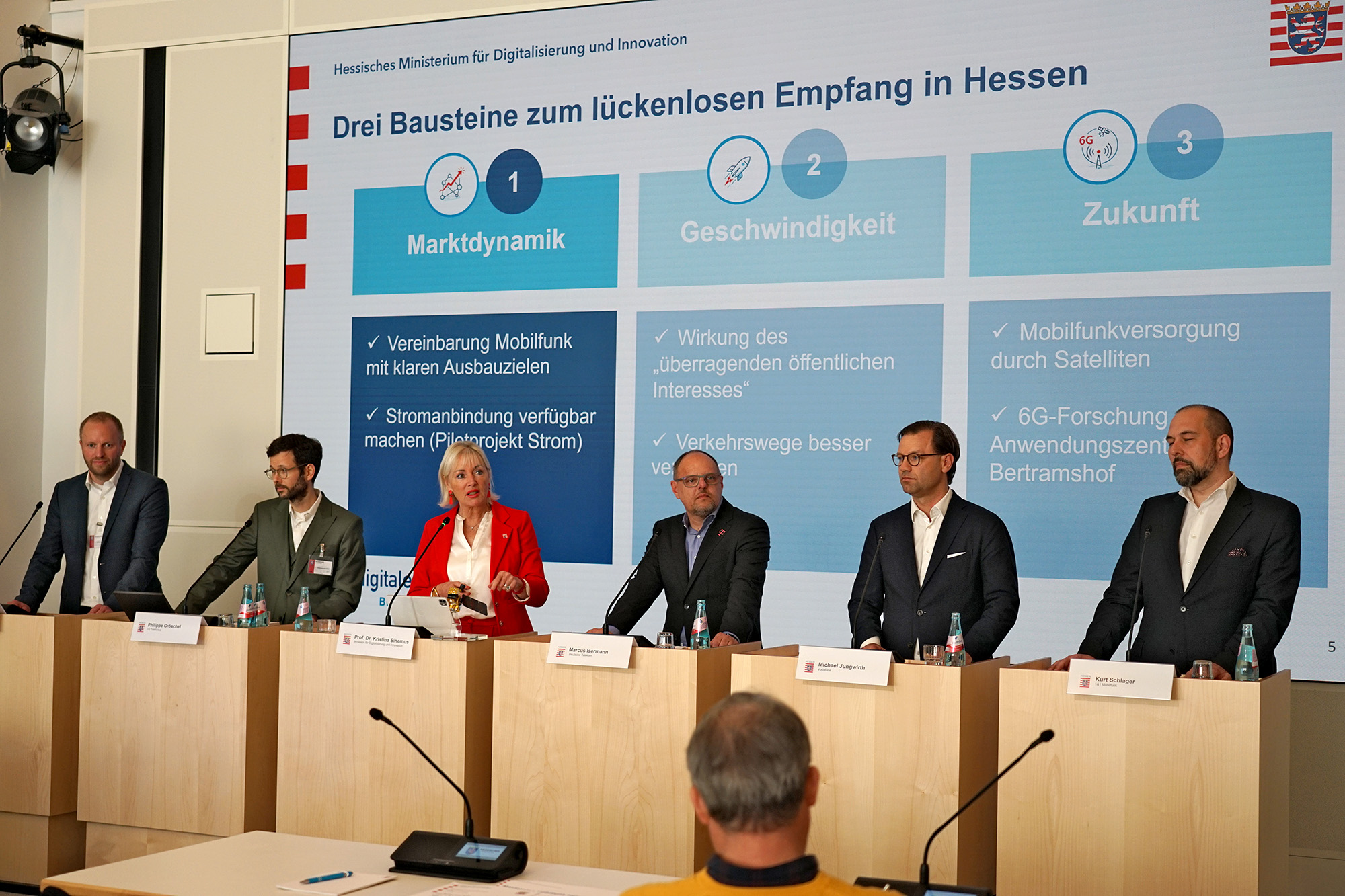 Sechs in einer Reihe stehende Rednerpulte, alle bis auf eines mit dunkel gekleideten Männern besetzt, am 3. von links Kristina Sinemus in leuchtend rotem Blazer.