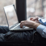 Beine und Torso eines Mannes mmit hellblauem Business-Hemd, der liegend auf einem Laptop tippt.