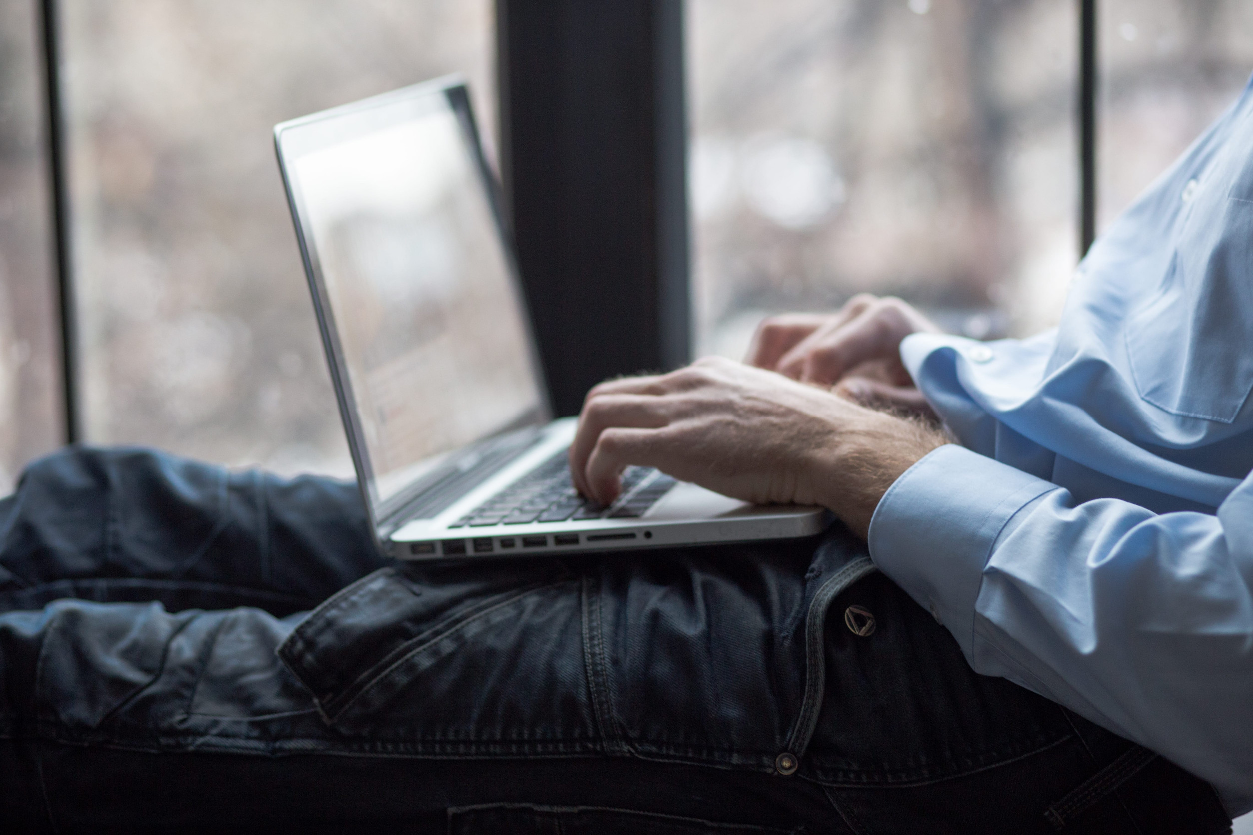 Beine und Torso eines Mannes mmit hellblauem Business-Hemd, der liegend auf einem Laptop tippt.