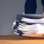 Ein paar offenbar männliche Hände sind dabei, einen Aktenstapel auf einer hölzernen Tischplatte abzulegen.