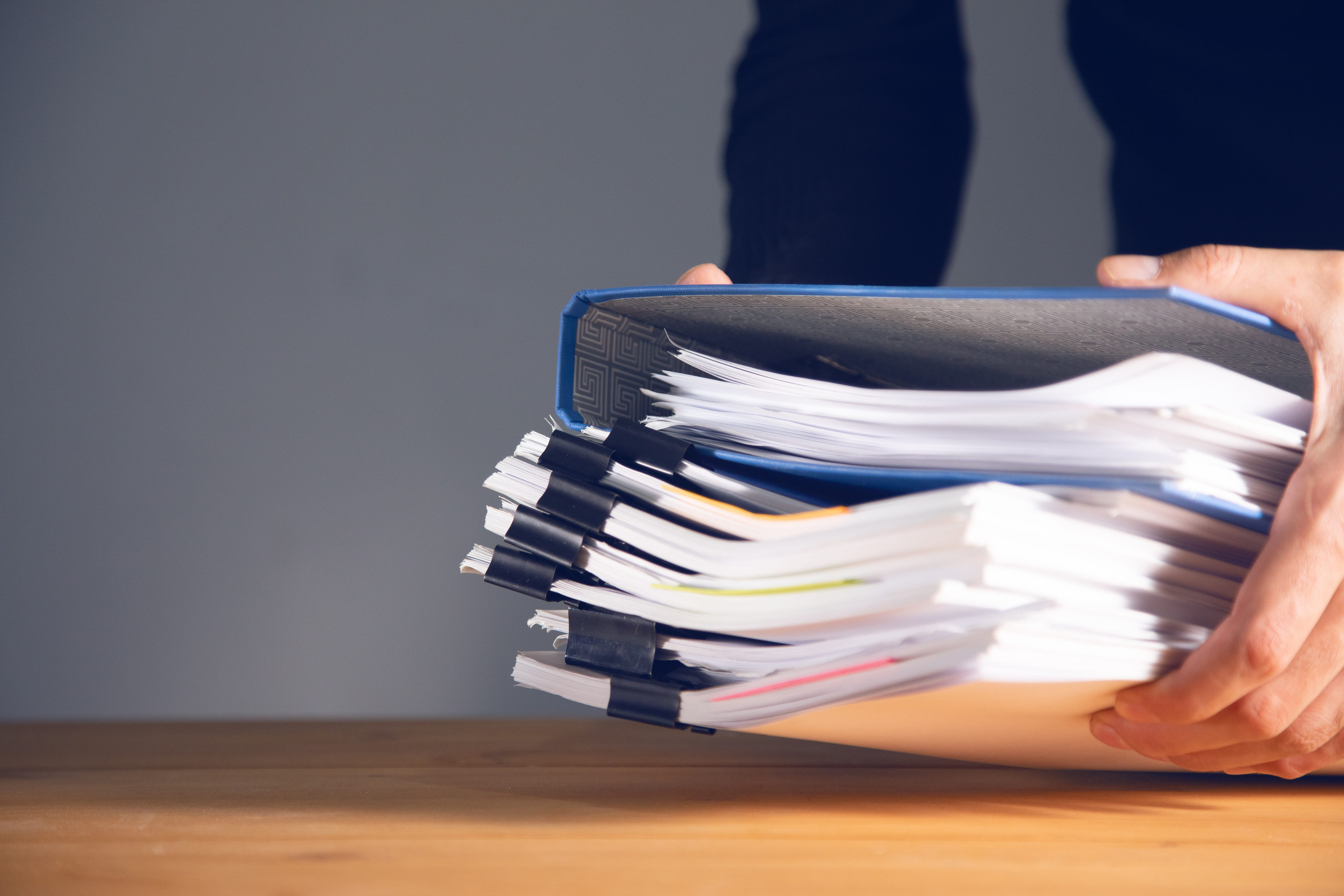 Ein paar offenbar männliche Hände sind dabei, einen Aktenstapel auf einer hölzernen Tischplatte abzulegen.
