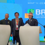 Vier Personen an zwei Podien, mit Dokumenten, die gerade unterschrieben werden. Im Hintergrund eine nicht erkennbare Projektion, im Vordergrund eine Deutschlandflagge im Anschnitt.