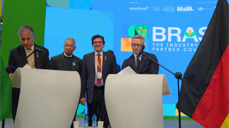 Vier Personen an zwei Podien, mit Dokumenten, die gerade unterschrieben werden. Im Hintergrund eine nicht erkennbare Projektion, im Vordergrund eine Deutschlandflagge im Anschnitt.