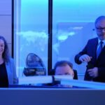 In blaues Licht getaquchter Raum, Person am Monitor, flankiert von Mann und Frau in förmlicher Kleidung