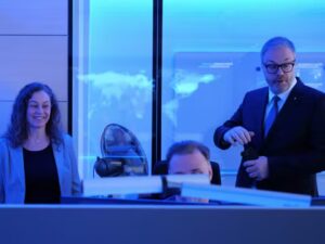 In blaues Licht getaquchter Raum, Person am Monitor, flankiert von Mann und Frau in förmlicher Kleidung