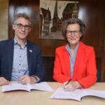 Ein Mann und einbe Frau in Business-Kleidung sitzen nebeneinander an einem Holztisch, vor sich haben sie Papiere, die sie gerade unterzeichnen.