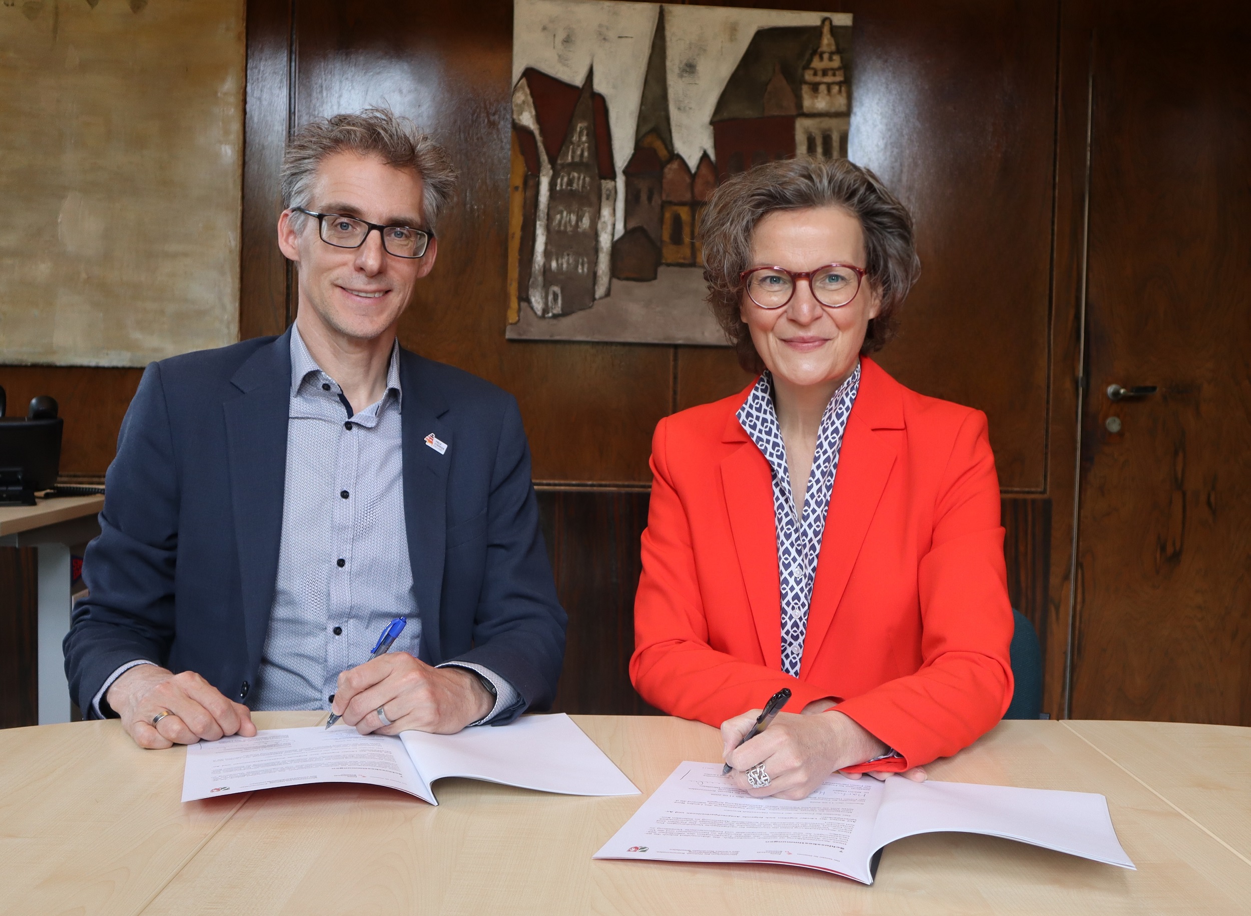 Ein Mann und einbe Frau in Business-Kleidung sitzen nebeneinander an einem Holztisch, vor sich haben sie Papiere, die sie gerade unterzeichnen.