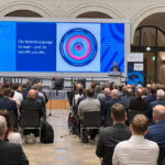 BSI-Präsidentin Claudia Plattner steht in einem blauen Sakko an einem Rednerpult, hinter ihr wird eine blaue Grafik auf einer Leinwand angezeigt. Vor ihr sitzt das Publikum auf Stühlen in einem Saal mit hohen Decken.