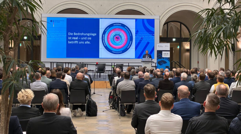BSI-Präsidentin Claudia Plattner steht in einem blauen Sakko an einem Rednerpult, hinter ihr wird eine blaue Grafik auf einer Leinwand angezeigt. Vor ihr sitzt das Publikum auf Stühlen in einem Saal mit hohen Decken.