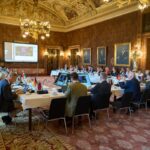 Großer Saal mit barocken Deckenelementen und rotem Teppich, darin eine U-förmige Tischanordnung und zahlreiche menschen in Anzügen und Kostümen sowie Monitore