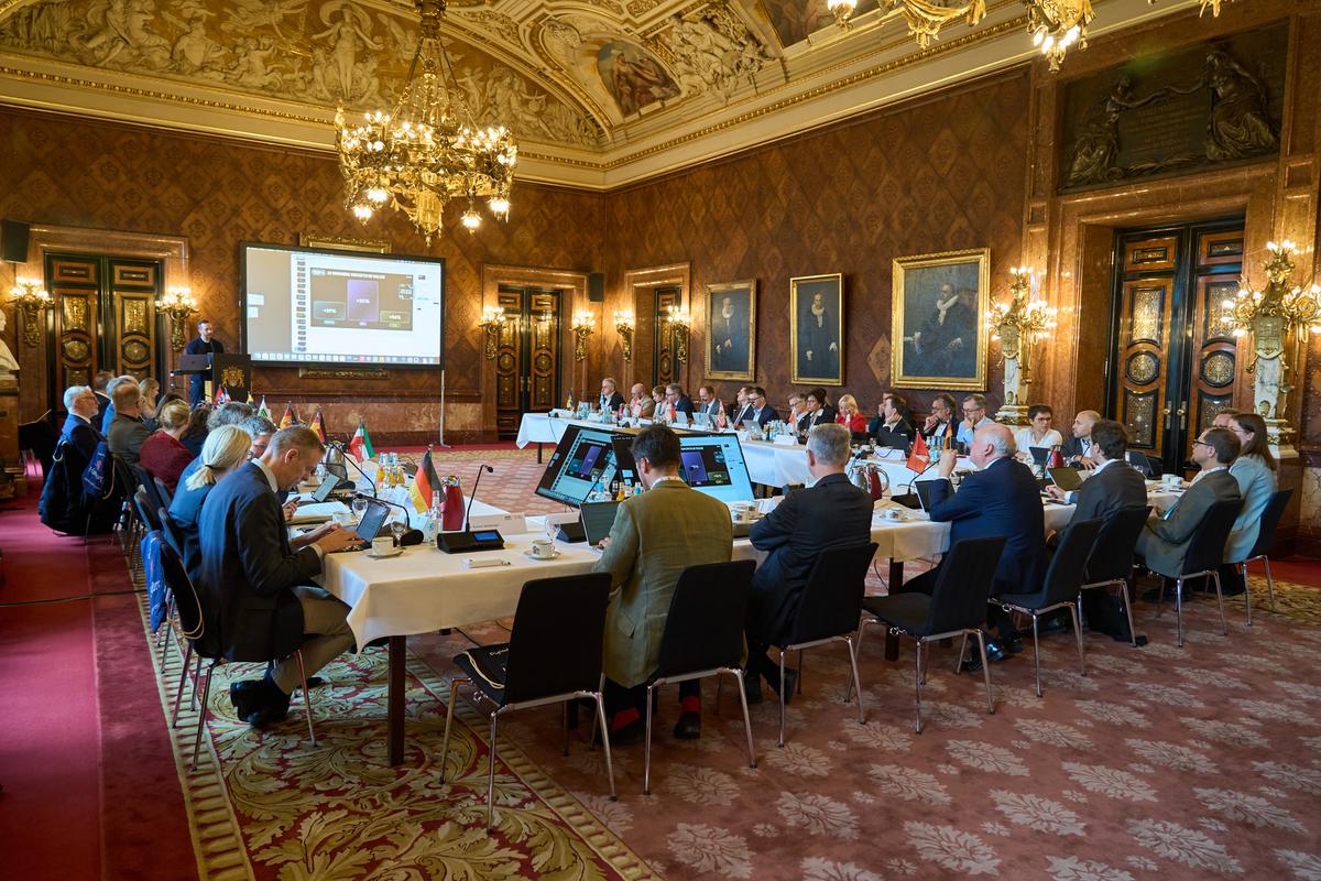 Großer Saal mit barocken Deckenelementen und rotem Teppich, darin eine U-förmige Tischanordnung und zahlreiche menschen in Anzügen und Kostümen sowie Monitore