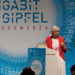 Ktistina Sinemus auf der Bühne beim Gigabitgipfel 2026; roter Blazer, cyanfarbene Bühne