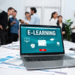 Nüchternes Büro mit einigen herumstehenden Personen im Hintergrud, vorne auf dem Tisch laptop mit E-Learning-Log-In-Screen