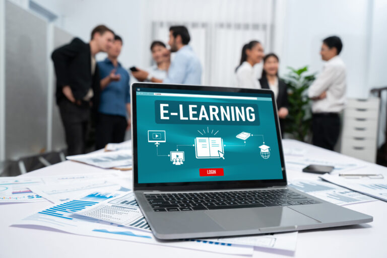 Nüchternes Büro mit einigen herumstehenden Personen im Hintergrud, vorne auf dem Tisch laptop mit E-Learning-Log-In-Screen