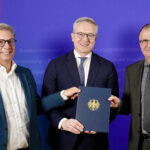 Karsten Wildberger steht vor einer violett-roten Wand zwischen zwei nicht näher benannten Männern, alle im Anzug
