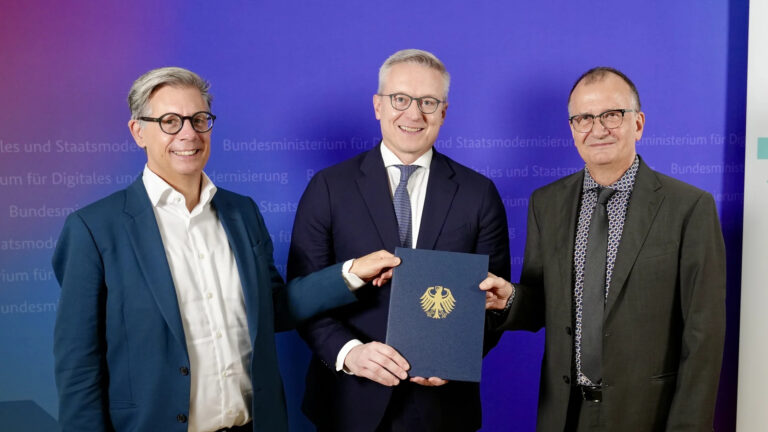 Karsten Wildberger steht vor einer violett-roten Wand zwischen zwei nicht näher benannten Männern, alle im Anzug