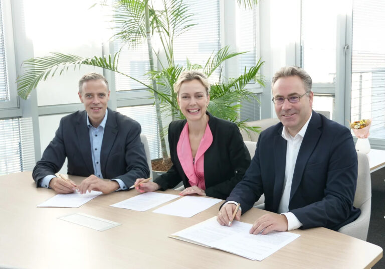 Heller Raum, heller Holztisch, zwei Männer und eine Frau sitzen nebeneinander mit Dokumenten vor sich und lächeln in die Kamera