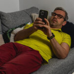 Mann mit gelbem Shirt und dunkelroter Hose liegt gemütlich auf einer grauen Couch und schaut auf sein Smartphone.