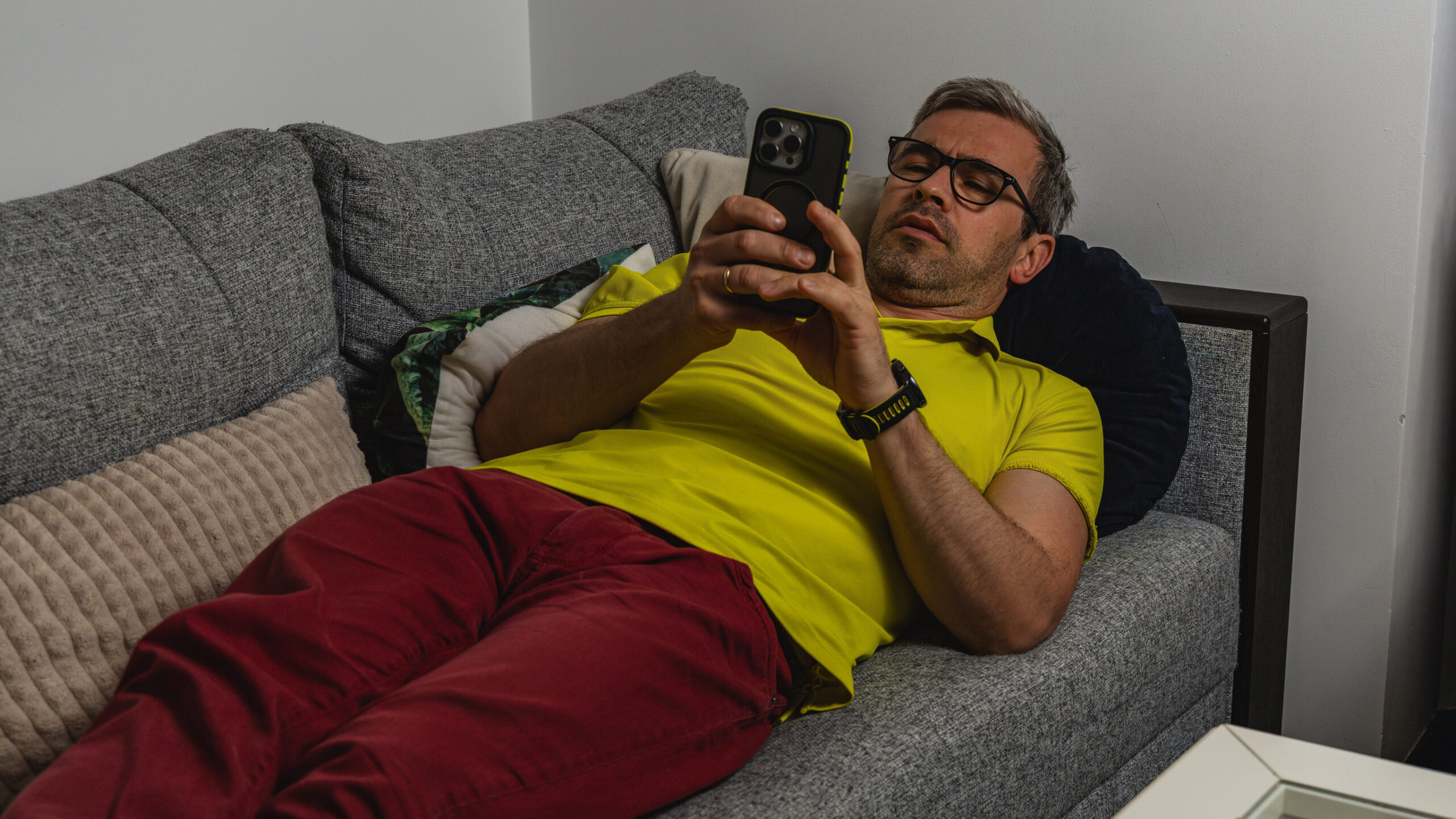 Mann mit gelbem Shirt und dunkelroter Hose liegt gemütlich auf einer grauen Couch und schaut auf sein Smartphone.