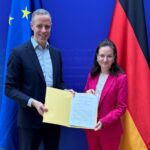 Staatssekretär Markus Richter und Chief Digital Officer Ronja Kemmer posieren mit einer Dokumnentenmappe vor blauem Hintergrund und Deutschland-Fahne