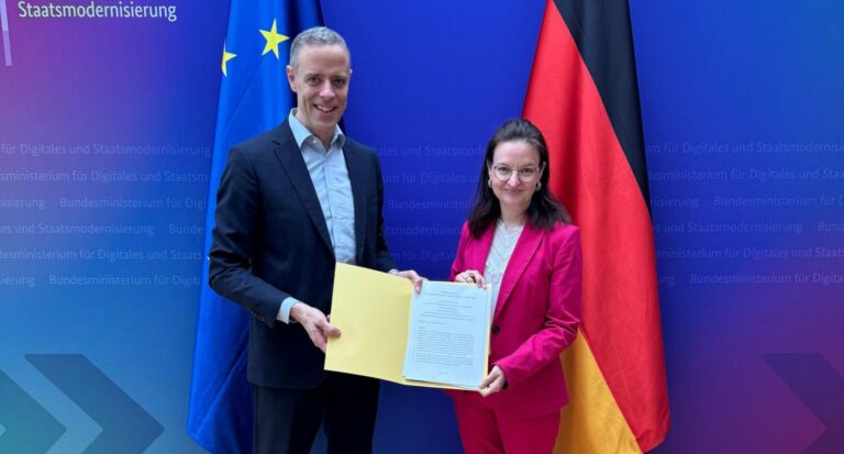 Staatssekretär Markus Richter und Chief Digital Officer Ronja Kemmer posieren mit einer Dokumnentenmappe vor blauem Hintergrund und Deutschland-Fahne